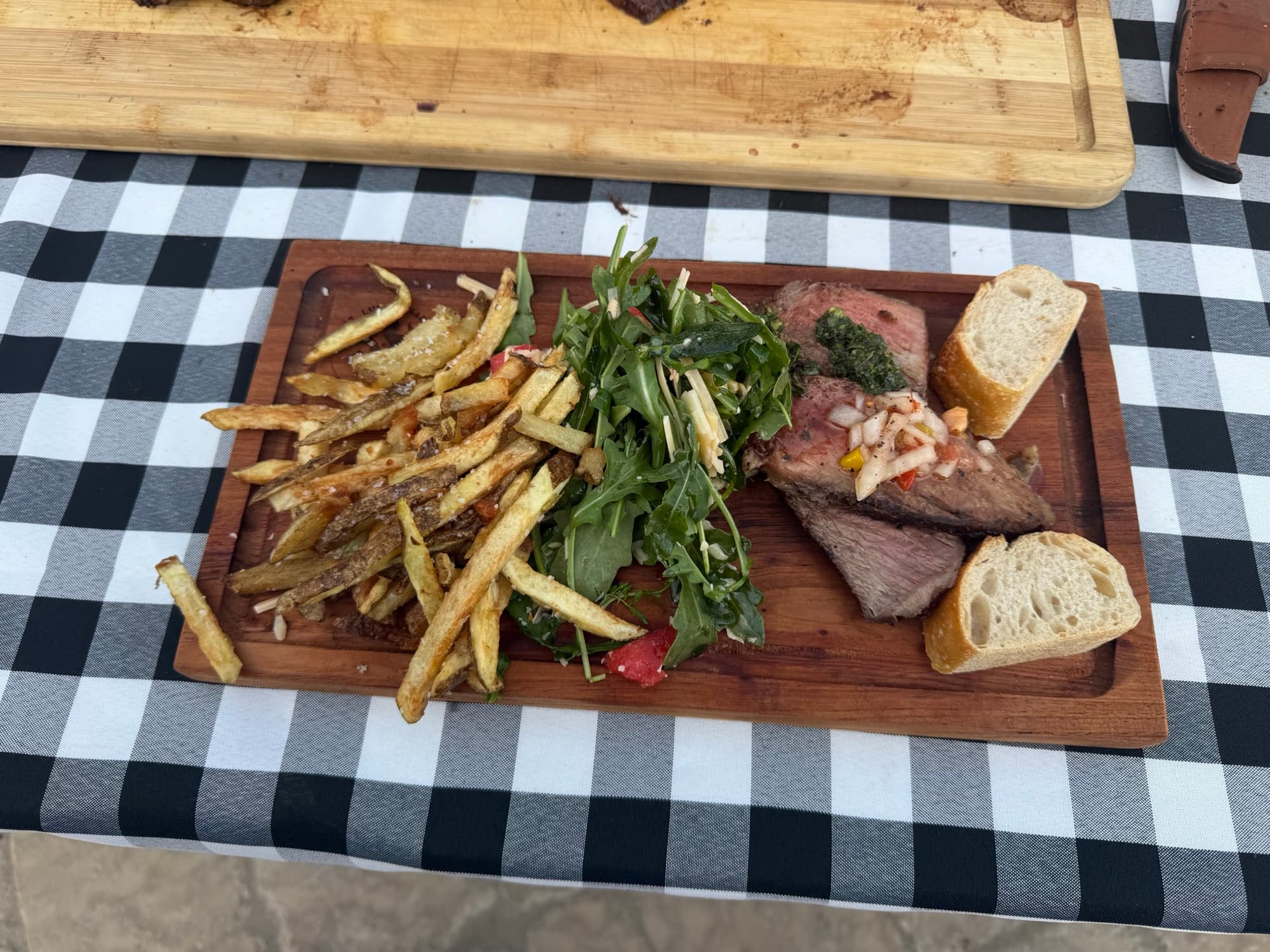 A wooden board plated with sliced beef, chimichurri, arugula salad, hand-cut fries, and crusty bread