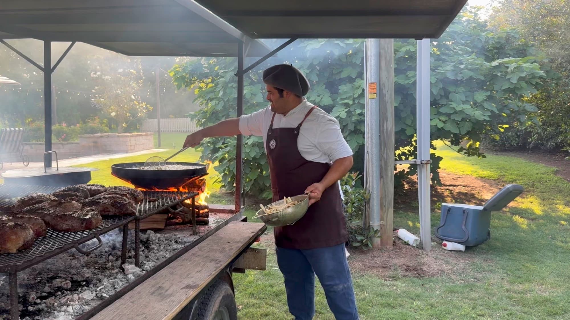 Pancho frying hand-cut potatoes in a pan over the open fire, meat resting on the grill beside him