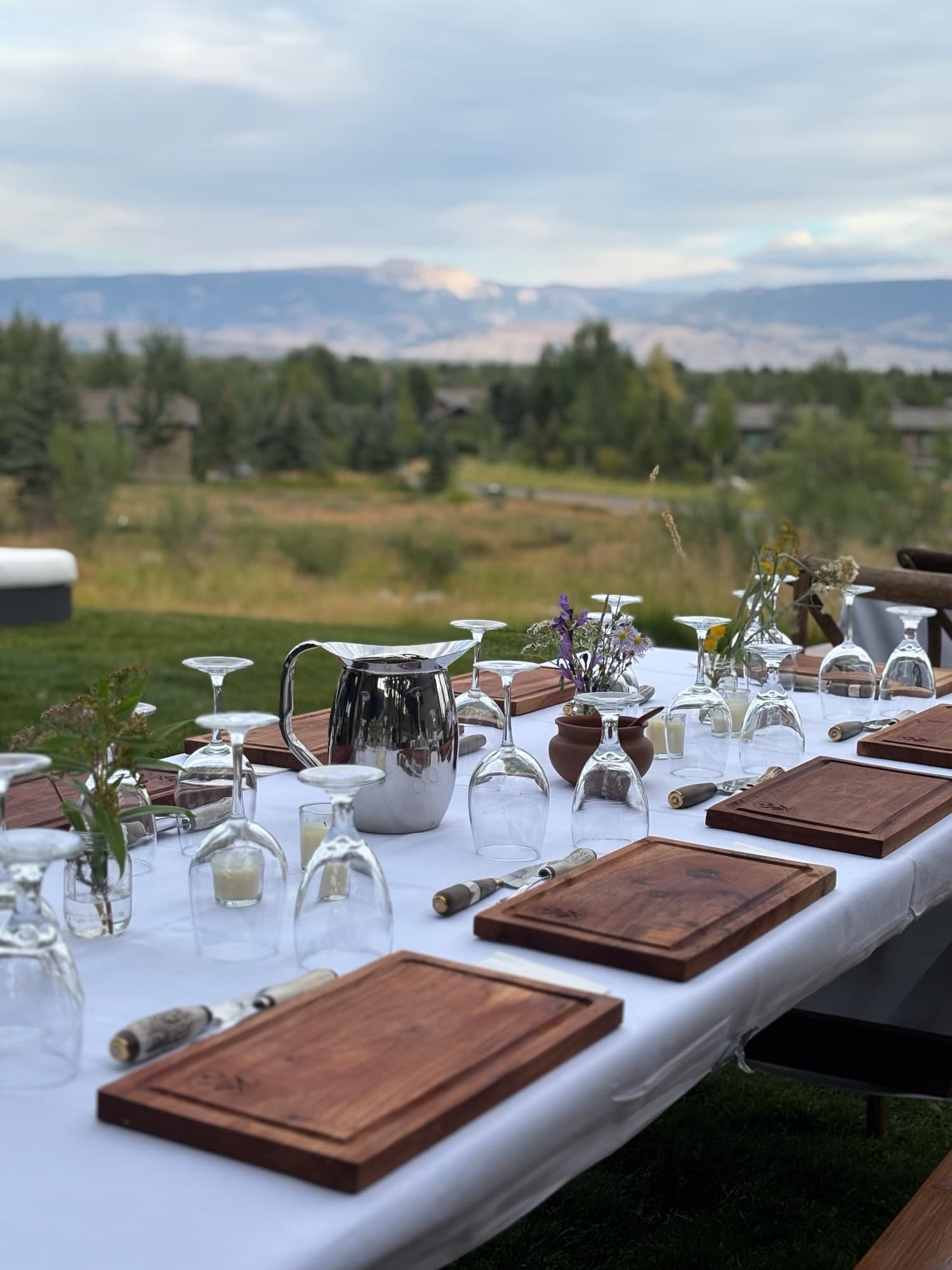 An outdoor table set with wooden boards, wildflowers, and glassware, a mountain range in the distance